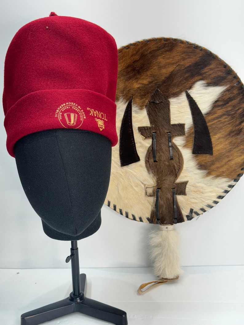 May include: A red wool beanie with the text "TONAK" and "MADE IN CZECH REPUBLIC" embroidered in gold. The beanie is on a black mannequin head. A brown and white cowhide shield with a leather strap is in the background.