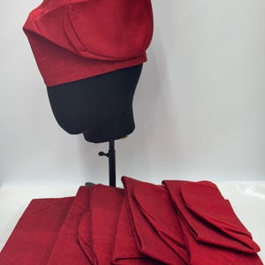 Può includere: Un set di dieci cappelli in tessuto rosso. I cappelli sono piegati e impilati uno sopra l'altro. I cappelli sono realizzati in un tessuto morbido e testurizzato.
