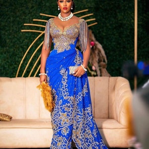 May include: A woman wearing a blue and gold beaded gown with a long train. She is standing in front of a white sofa and a green wall.