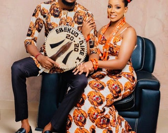 Könnte beinhalten: Ein Paar in traditioneller nigerianischer Kleidung. Die Frau trägt ein weißes und orangefarbenes Kleid mit einem Löwenmuster. Der Mann trägt ein weißes und braunes Hemd mit einem Löwenmuster und eine schwarze Hose. Sie halten ein rundes, braunes Objekt mit dem Text "EBEKUD 20.20.20. DIKE I" darauf.