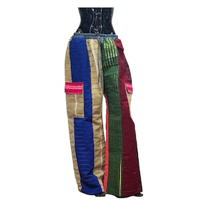 May include: Colorful patchwork pants with a drawstring waist. The pants feature a mix of blue, beige, green, and red panels with a textured, perforated design. Two pockets are visible, one pink and one red.