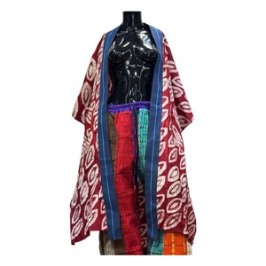 Op de afbeelding: Een rode kimono-achtige mantel met een wit bladerpatroon, blauwe bies en een veelkleurige patchwork rok. De mantel is open en onthult de outfit eronder. De outfit wordt getoond op een zwarte mannequin.