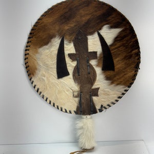 May include: A round, brown and white cowhide fan with a black leather design in the center. The fan has a white fur handle with a brown leather strap.