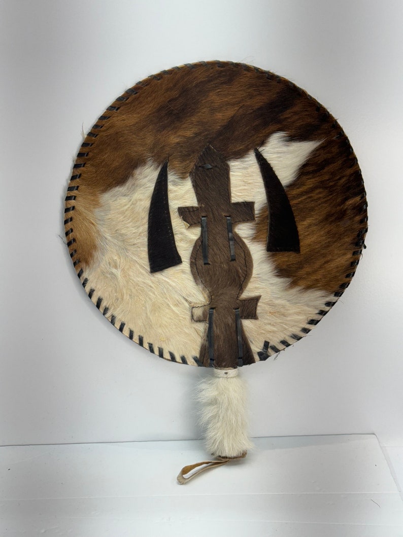 May include: A round, brown and white cowhide fan with a black leather design in the center. The fan has a white fur handle with a brown leather strap.
