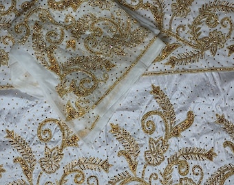 Puede incluir: Una tela blanca con intrincados bordados dorados y lentejuelas doradas dispersas. El diseño consiste en patrones florales, motivos de hojas y volutas ornamentadas. La tela está doblada, mostrando una vista en capas de los adornos.