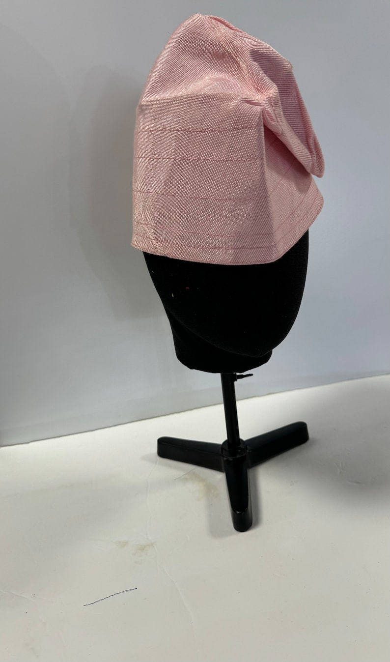 May include: A pink fabric hat with a textured pattern. The hat is on a black mannequin head.