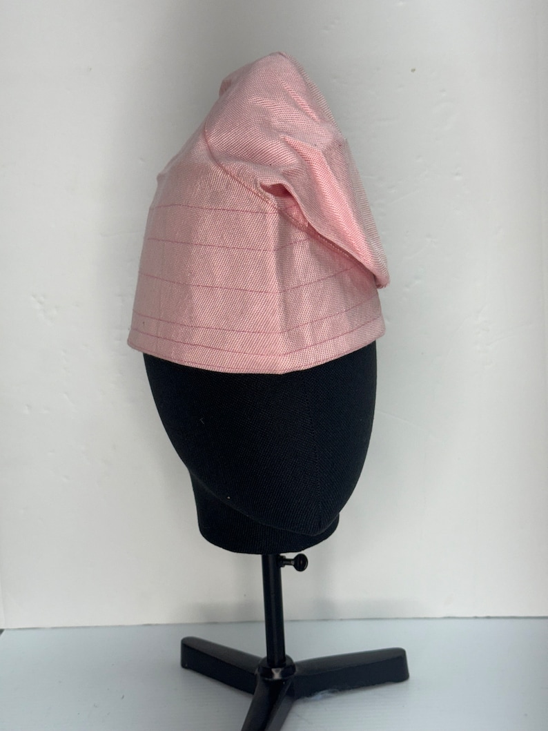 May include: A pink, patterned fabric hat with a pointed top. The hat is on a black mannequin head.