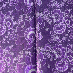 Puede incluir: Primer plano de una tela con un estampado floral. El diseño presenta flores grandes y pequeñas en tonos morados y blancos sobre un fondo morado oscuro. La tela tiene una apariencia texturizada.