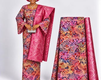 Könnte beinhalten: Ein langärmeliges Kleid mit gewelltem Ausschnitt und Saum, das ein lebhaftes Blumenmuster in Rosa-, Orange- und Blautönen aufweist. Ein passendes, einfarbiges rosa Tuch mit strukturiertem Paisleymuster und eine große kreisförmige Kopfbedeckung.