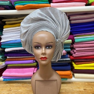 May include: A silver headwrap with a pleated design, worn on a mannequin head. The headwrap is made of a shiny fabric and has a unique, twisted shape.