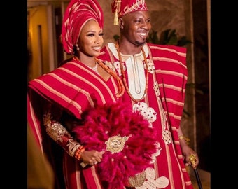 Puede incluir: Una pareja con atuendo tradicional de boda nigeriana. La mujer lleva una falda de rayas rojas y doradas con un turbante a juego. Lleva un gran abanico de plumas rosas. El hombre lleva una túnica de rayas rojas y doradas con un gorro a juego. Lleva un bastón.