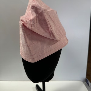 May include: A pink ribbed fabric bonnet sits on a black mannequin. The bonnet has a unique shape, with a pointed crown and a slightly curved brim. The fabric appears soft and smooth, and the overall style is simple and elegant.  The bonnet is displayed on a simple black stand against a white background. size 23