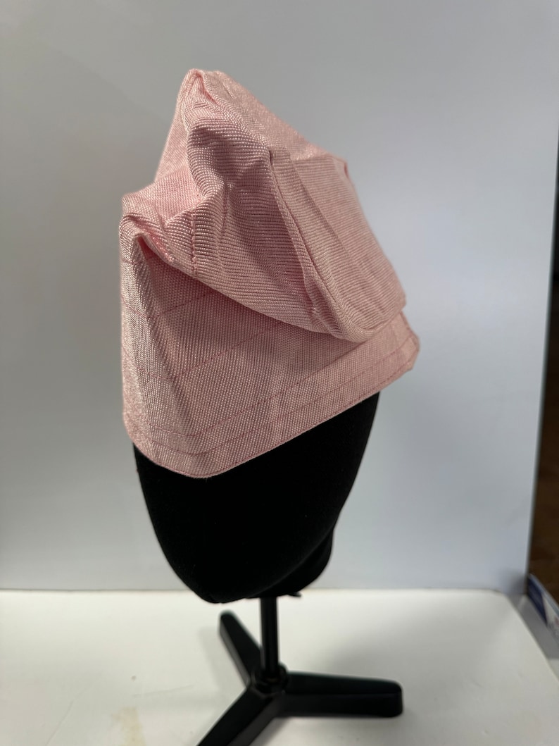 May include: A pink ribbed fabric bonnet sits on a black mannequin. The bonnet has a unique shape, with a pointed crown and a slightly curved brim. The fabric appears soft and smooth, and the overall style is simple and elegant.  The bonnet is displayed on a simple black stand against a white background. size 23