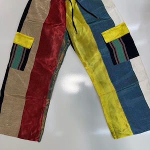 May include: Colorful patchwork pants with a drawstring waist. The pants feature vertical stripes in various colors, including black, tan, red, yellow, blue, and white. Two pockets with yellow flaps and patterned fabric accents are visible.