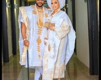 Puede incluir: Una pareja vestida con atuendos tradicionales de boda nigerianos blancos y dorados. La mujer lleva un turbante blanco y un vestido blanco con bordados dorados. El hombre lleva una túnica blanca con bordados dorados y una gorra blanca. Están de pie en un pasillo.