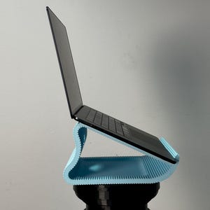 Pu&ograve; includere: Un supporto per laptop a forma di S di colore azzurro con una superficie scanalata e testurizzata che sostiene un laptop nero. Il supporto presenta un design curvo che solleva il dispositivo ad angolo, offrendo una base stabile.
