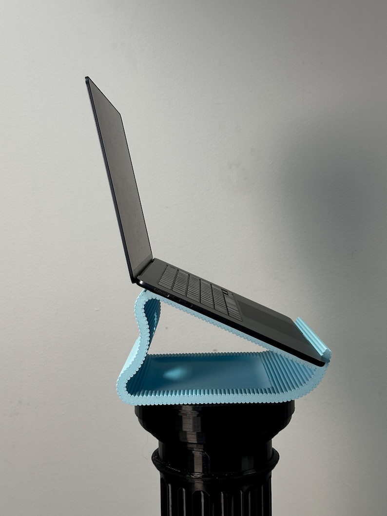 Pu&ograve; includere: Un supporto per laptop a forma di S di colore azzurro con una superficie scanalata e testurizzata che sostiene un laptop nero. Il supporto presenta un design curvo che solleva il dispositivo ad angolo, offrendo una base stabile.