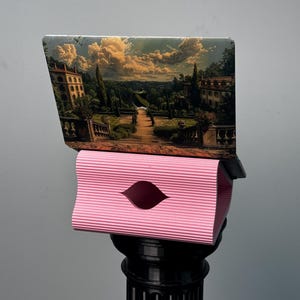 Pu&ograve; includere: Un computer portatile con una skin paesaggistica raffigurante un giardino e una villa, poggiato su un supporto rosa rettangolare a coste orizzontali con un'apertura centrale a mandorla, su una base a colonna nera scanalata.