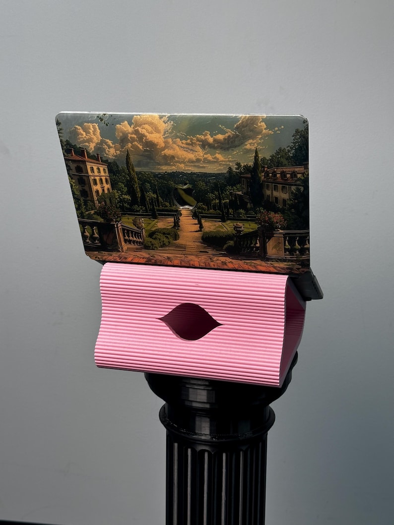 Pu&ograve; includere: Un computer portatile con una skin paesaggistica raffigurante un giardino e una villa, poggiato su un supporto rosa rettangolare a coste orizzontali con un'apertura centrale a mandorla, su una base a colonna nera scanalata.