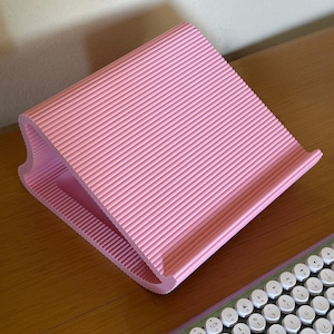 Pu&ograve; includere: Un supporto per laptop rosa chiaro caratterizzato da una superficie rigata e testurizzata con un profilo laterale curvo. Il supporto &egrave; angolato per sostenere un dispositivo ed &egrave; posizionato accanto a una parte di una tastiera con tasti bianchi.