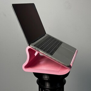 Pu&ograve; includere: Un supporto per laptop rosa chiaro, ondulato e stampato in 3D con una consistenza a coste che sostiene un laptop argentato. Il supporto presenta una forma curva ed ergonomica progettata per sollevare il dispositivo con un'angolazione.