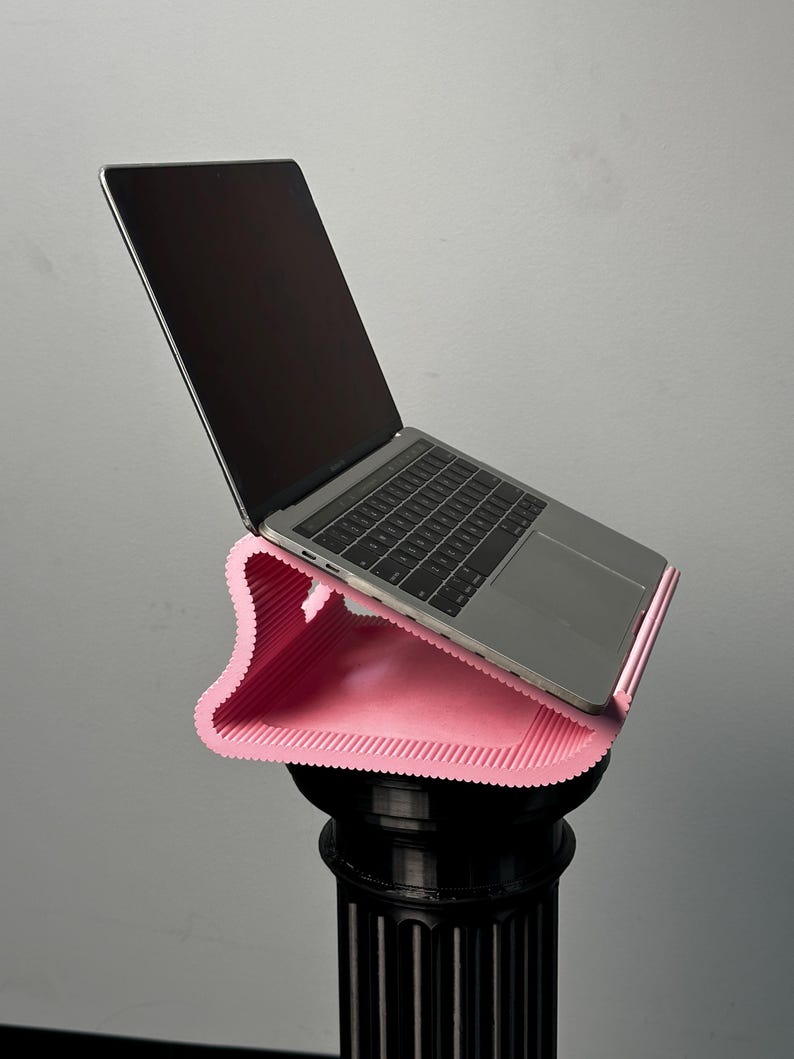 Pu&ograve; includere: Un supporto per laptop rosa chiaro, ondulato e stampato in 3D con una consistenza a coste che sostiene un laptop argentato. Il supporto presenta una forma curva ed ergonomica progettata per sollevare il dispositivo con un'angolazione.