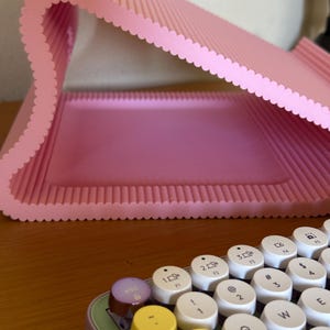 Pu&ograve; includere: Un supporto per portatile rosa chiaro a forma di S con una texture a coste e bordi smerlati. In primo piano si trova una parte di una tastiera con tasti rotondi bianchi con lettere nere, un tasto escape viola e tasti tab e blocco gialli.