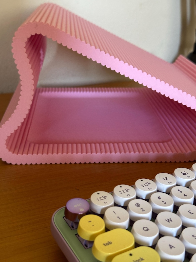 Pu&ograve; includere: Un supporto per portatile rosa chiaro a forma di S con una texture a coste e bordi smerlati. In primo piano si trova una parte di una tastiera con tasti rotondi bianchi con lettere nere, un tasto escape viola e tasti tab e blocco gialli.
