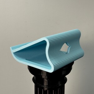 Pu&ograve; includere: Un supporto azzurro stampato in 3D con una texture ondulata e nervata e un profilo triangolare. L'oggetto presenta un ritaglio centrale a forma di diamante sul lato e poggia su un piedistallo cilindrico nero con scanalature verticali.