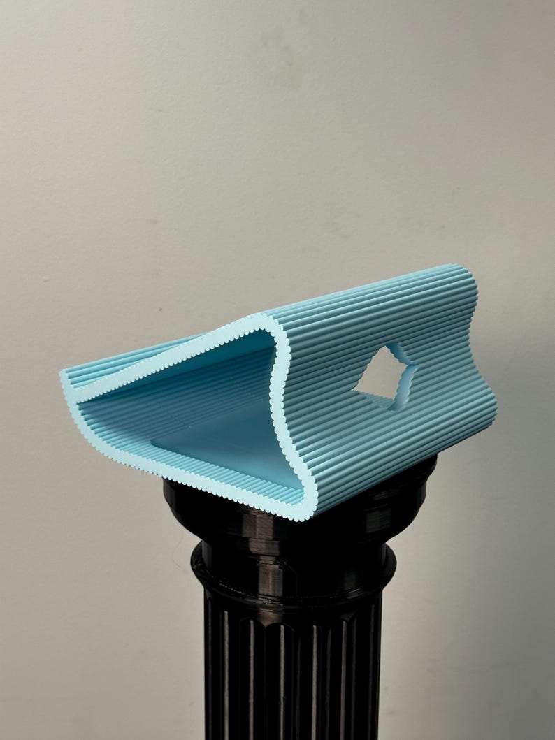 Pu&ograve; includere: Un supporto azzurro stampato in 3D con una texture ondulata e nervata e un profilo triangolare. L'oggetto presenta un ritaglio centrale a forma di diamante sul lato e poggia su un piedistallo cilindrico nero con scanalature verticali.