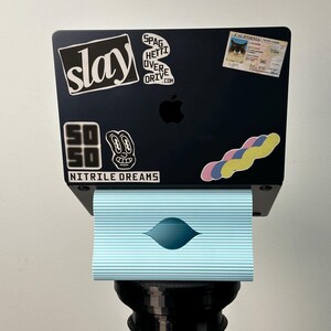 Pu&ograve; includere: Un portatile nero con vari adesivi tra cui il testo slay, SPAGHETTI OVERDRIVE.COM, SO SO NITRILE DREAMS e una patente di guida della California, poggiato su un supporto azzurro a coste orizzontali sopra un piedistallo nero.