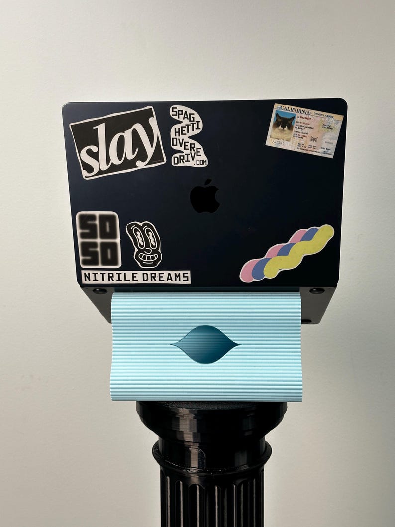Pu&ograve; includere: Un portatile nero con vari adesivi tra cui il testo slay, SPAGHETTI OVERDRIVE.COM, SO SO NITRILE DREAMS e una patente di guida della California, poggiato su un supporto azzurro a coste orizzontali sopra un piedistallo nero.
