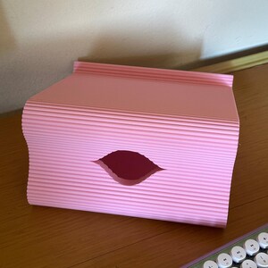 Pu&ograve; includere: Un organizer da scrivania rettangolare rosa chiaro caratterizzato da una texture a coste e ondulata sulle superfici esterne. Il pannello frontale presenta un'apertura centrale a forma di mandorla che rivela lo spazio interno.