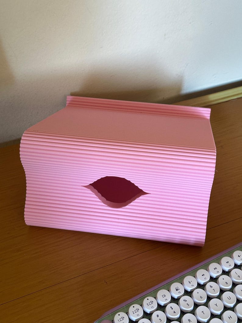 Pu&ograve; includere: Un organizer da scrivania rettangolare rosa chiaro caratterizzato da una texture a coste e ondulata sulle superfici esterne. Il pannello frontale presenta un'apertura centrale a forma di mandorla che rivela lo spazio interno.