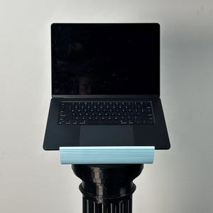 Pu&ograve; includere: Un computer portatile nero poggia su un supporto azzurro a coste orizzontali, sostenuto da un alto piedistallo cilindrico nero scanalato. Lo schermo del portatile &egrave; scuro e la tastiera presenta una disposizione standard dei tasti QWERTY.