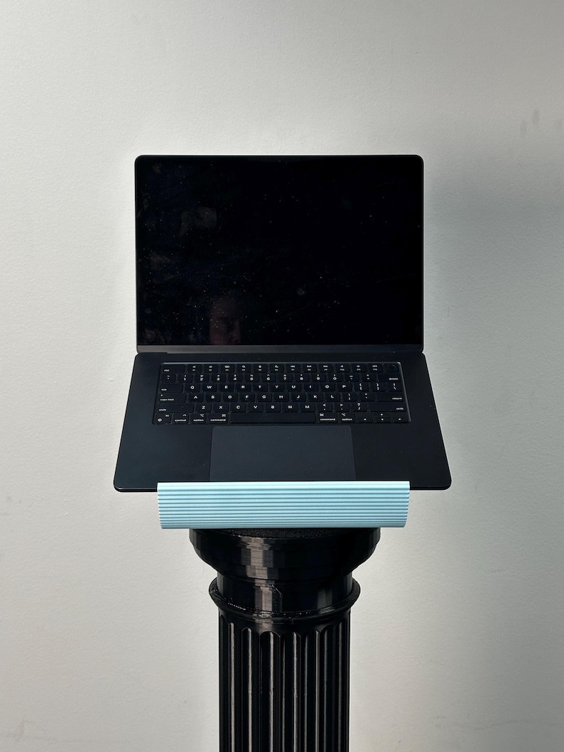 Pu&ograve; includere: Un computer portatile nero poggia su un supporto azzurro a coste orizzontali, sostenuto da un alto piedistallo cilindrico nero scanalato. Lo schermo del portatile &egrave; scuro e la tastiera presenta una disposizione standard dei tasti QWERTY.