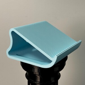 Pu&ograve; includere: Un supporto a forma di cuneo, stampato in 3D, di colore azzurro con una superficie scanalata e testurizzata. L'oggetto presenta un design curvo con lati aperti, una base piatta e una superficie superiore inclinata su un piedistallo nero.
