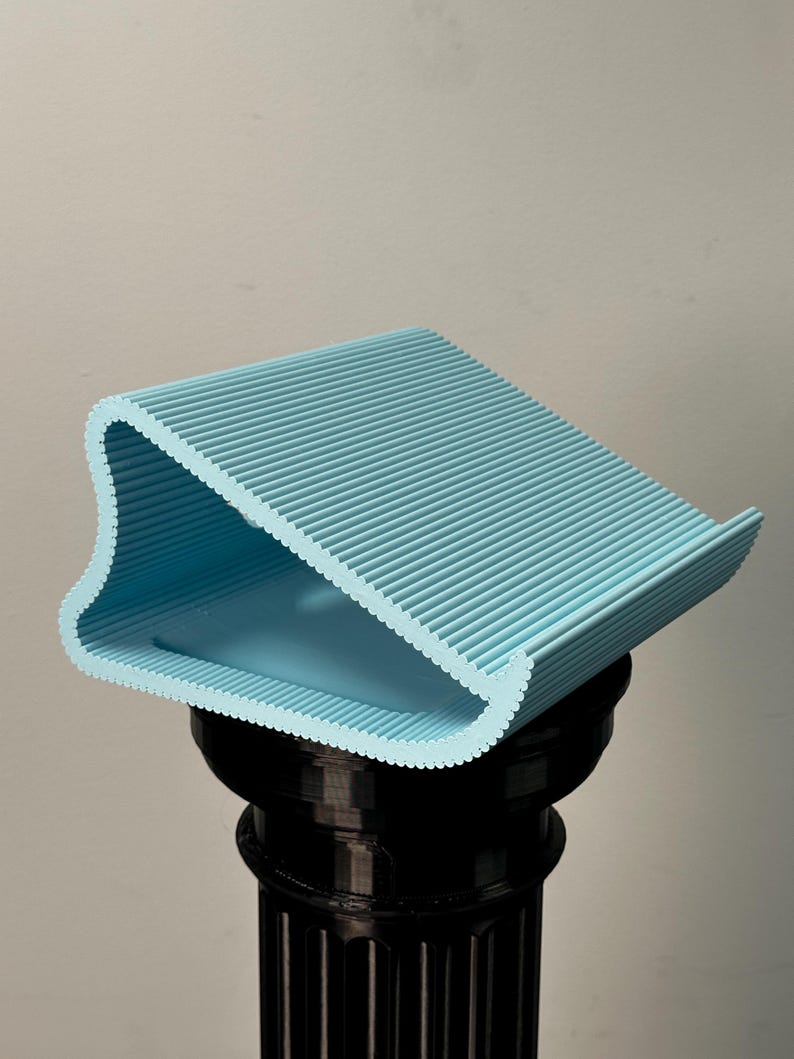 Pu&ograve; includere: Un supporto a forma di cuneo, stampato in 3D, di colore azzurro con una superficie scanalata e testurizzata. L'oggetto presenta un design curvo con lati aperti, una base piatta e una superficie superiore inclinata su un piedistallo nero.