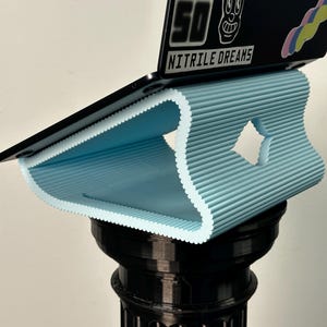 Pu&ograve; includere: Un supporto per laptop azzurro, a coste e curvo con un ritaglio a forma di diamante, che sostiene un coperchio nero per laptop con un adesivo recante il testo "50 50" e "NITRILE DREAMS" sopra un elemento grafico colorato e ondulato.