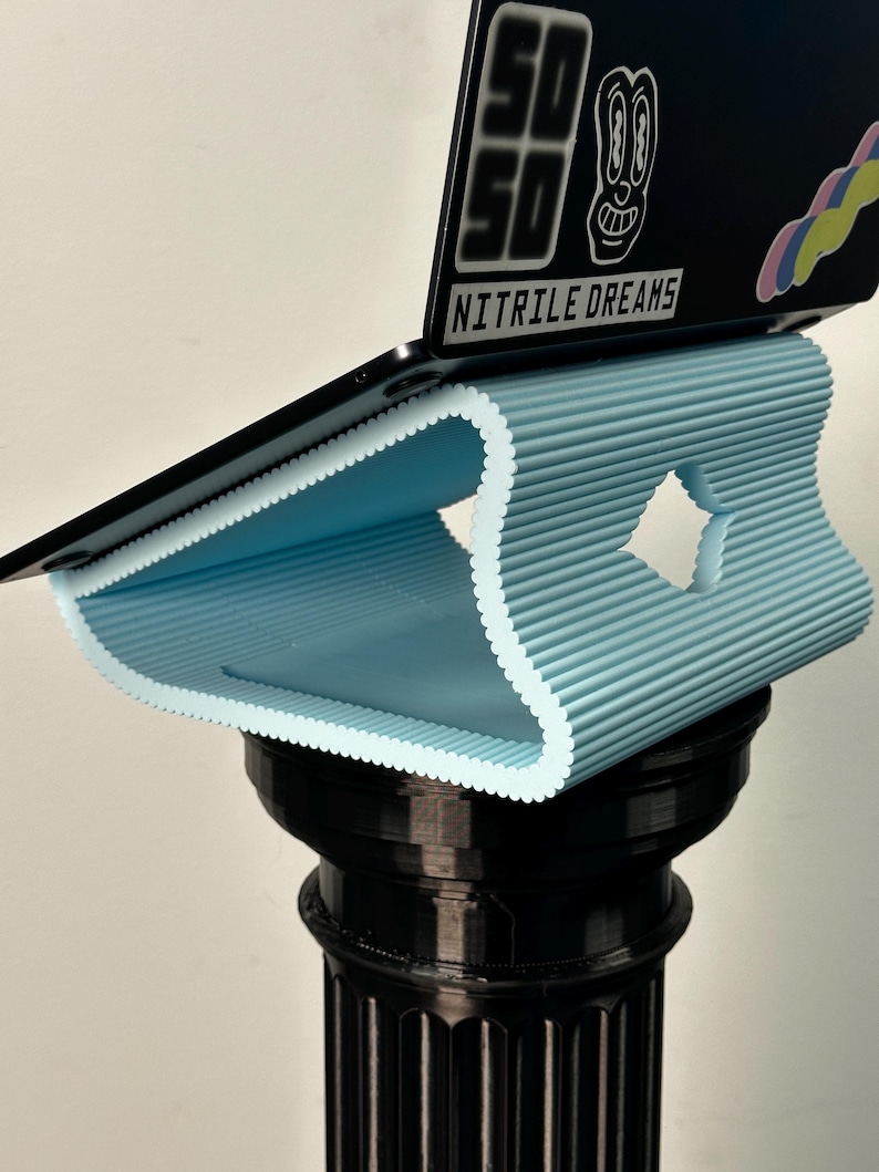 Pu&ograve; includere: Un supporto per laptop azzurro, a coste e curvo con un ritaglio a forma di diamante, che sostiene un coperchio nero per laptop con un adesivo recante il testo "50 50" e "NITRILE DREAMS" sopra un elemento grafico colorato e ondulato.