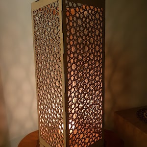 Peut inclure: Une lampe en bois avec un motif géométrique découpé. La lampe est allumée et diffuse une lueur chaude.