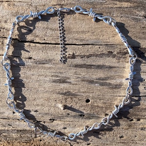 Puede incluir: Un collar de alambre de púas plateado con cierre de cadena. El collar está hecho de alambre retorcido con puntas afiladas.