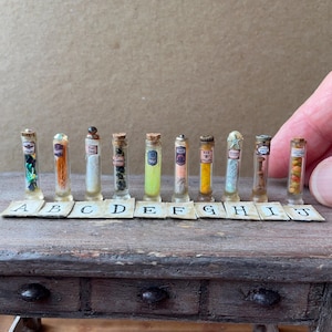 Peut inclure: Dix fioles en verre miniatures alignées, contenant chacune des matériaux de couleurs différentes et étiquetées avec une lettre de A à J. Les fioles sont dotées de bouchons en liège ou en métal et contiennent des herbes, poudres et perles.