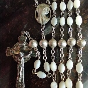 May include: A silver rosary with white pearl beads and a silver crucifix with a figure of Jesus Christ. The rosary also features a silver pendant with a depiction of the Virgin Mary.