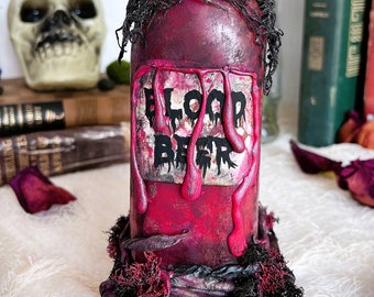 May include: A decorative bottle with a dark red finish and the words "BLOOD BEER" on a label. The bottle is adorned with black and red accents, moss, and dripping red paint, creating a spooky, gothic aesthetic. A skull and dried roses are in the background.