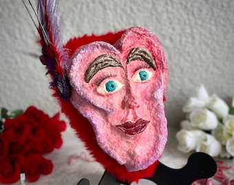 Könnte beinhalten: Eine rosafarbene herzförmige Skulptur mit einem menschlichen Gesicht, blauen Augen und roten Lippen. Die Skulptur ist mit einer lila Feder verziert und steht auf einem schwarzen Sockel.