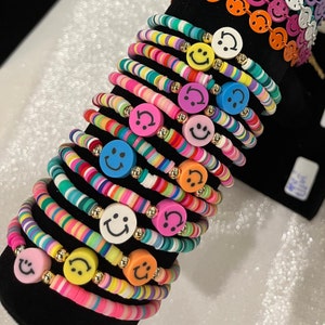 Puede incluir: Una pila de pulseras coloridas con dijes de caritas sonrientes. Cada pulsera está hecha de pequeñas cuentas coloridas y presenta un dije de carita sonriente de color diferente.