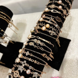 Puede incluir: Un expositor de pulseras de cadena dorada con detalles de perlas y diversos encantos, incluyendo caras sonrientes, estrellas, corazones y un ojo. Las pulseras se exhiben en un soporte negro.