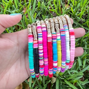 Puede incluir: Un conjunto de pulseras coloridas hechas con cuentas de plástico redondas apiladas. Las pulseras están enhebradas en cadenas de metal de color dorado.