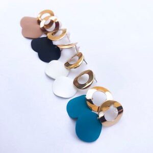 Puede incluir: Un conjunto de seis pares de pendientes con aros dorados y respaldos en forma de corazón de diferentes colores. Los colores incluyen marrón, negro, blanco y azul.